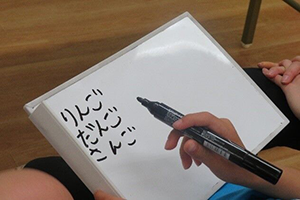 写真：手元のホワイドボードにひらがなを書く。「りんご」「だんご」「さんご」
